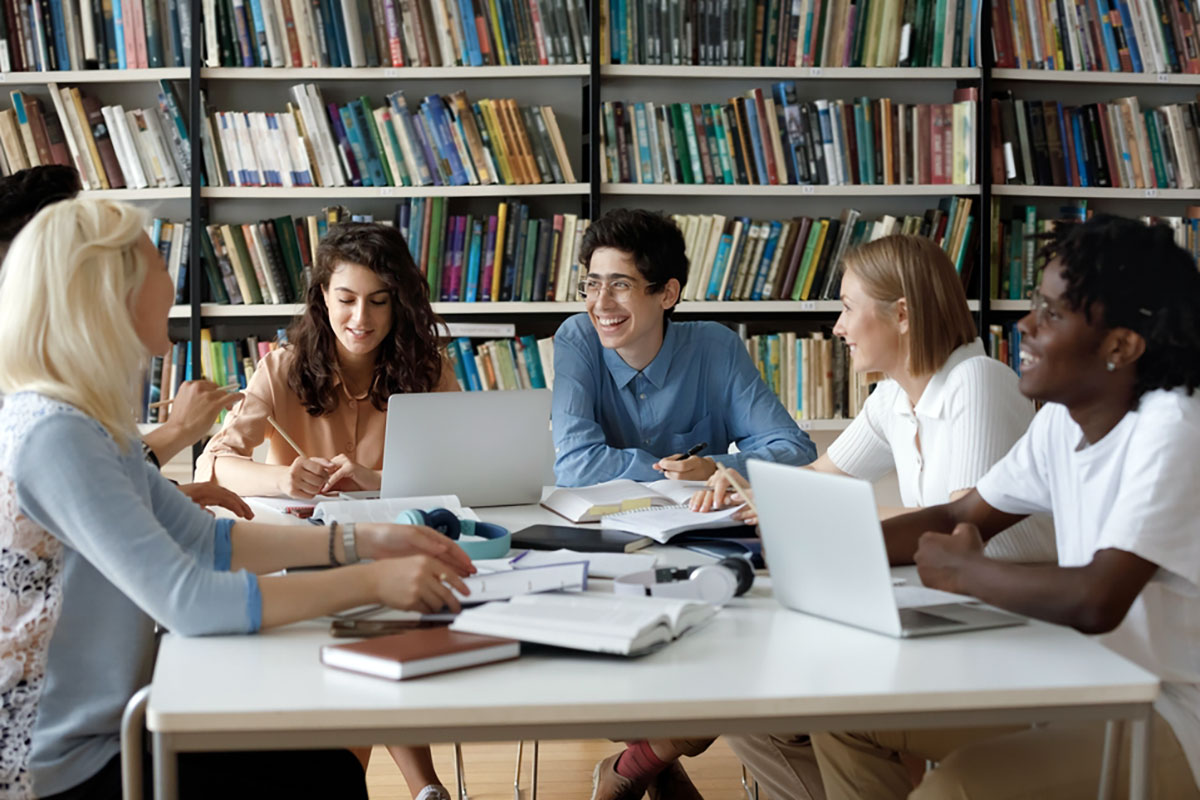 Five young people in conference at a table covered with books and laptops, in front of a wall of books on shelves
